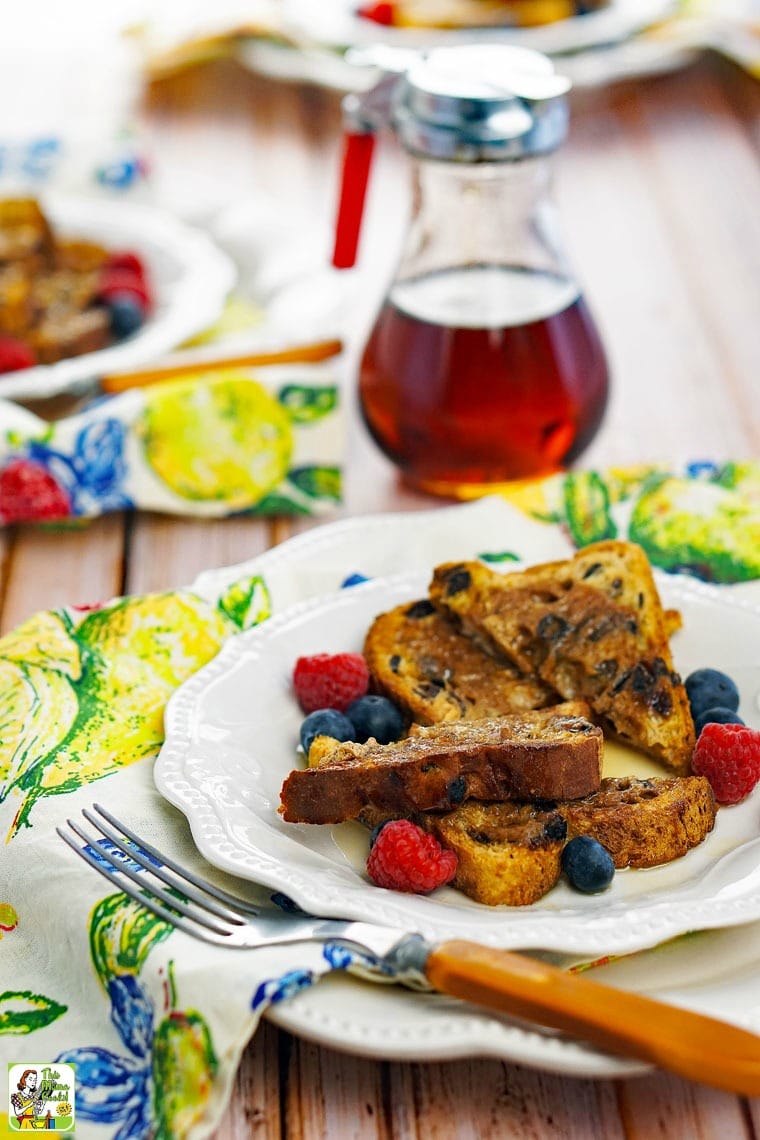 A white plate of French Toast with berries and yelllow fork a glass pitcher of maple syrup