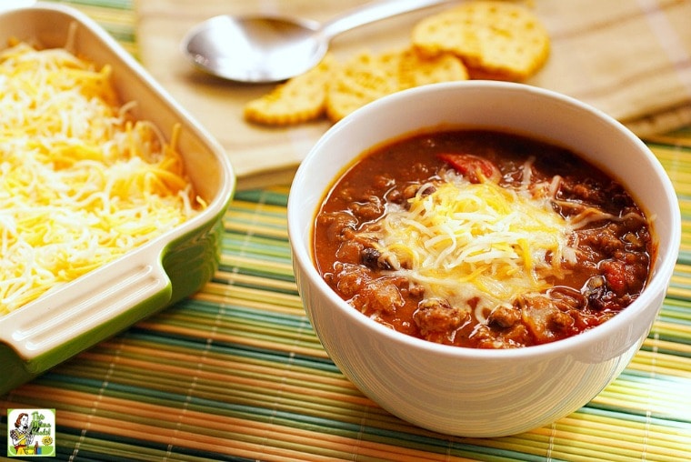 A bowl of Slow Cooker Pumpkin Chili with a container of shredded cheese, a wooden cutting board, crackers, and a wooden cutting board all on a bamboo mat.