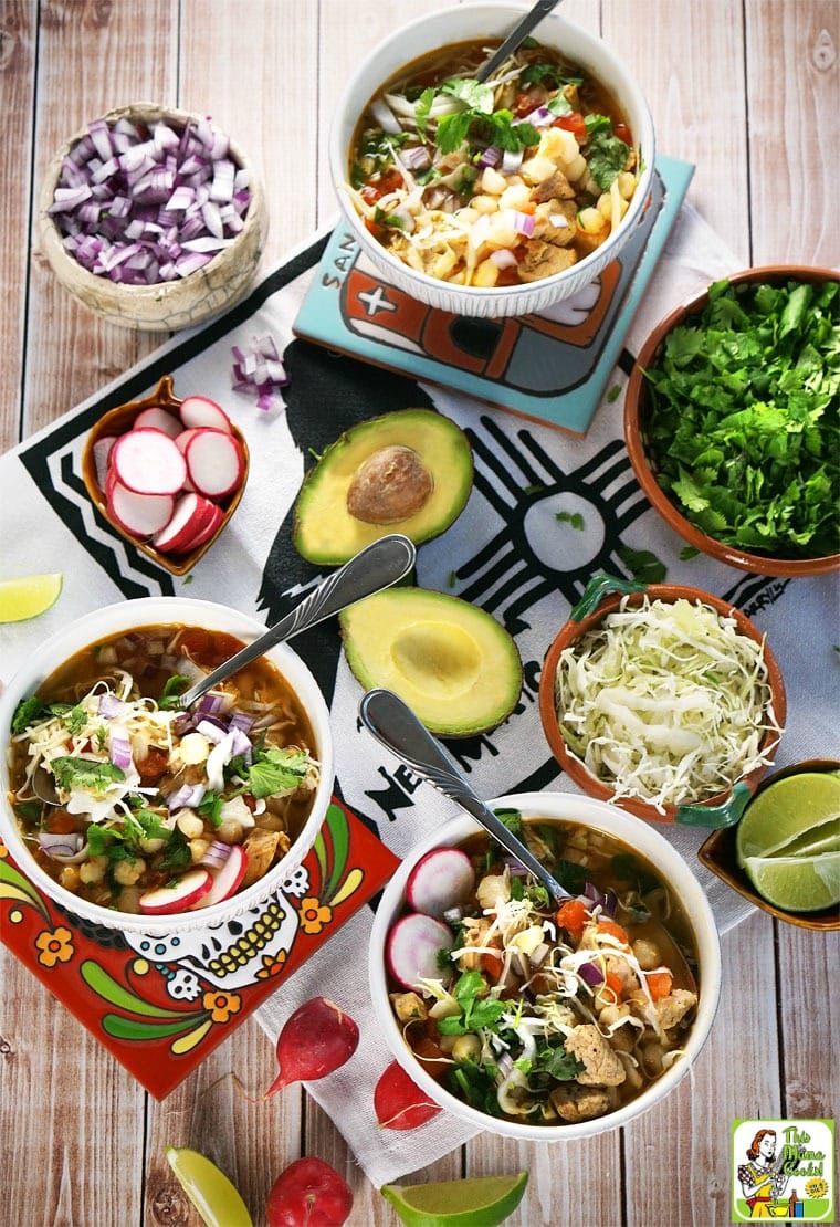 Bowls of pozole soup with bowls of toppings like avocado, shredded cabbage, cilantro, diced onions, diced onions, and limes. 