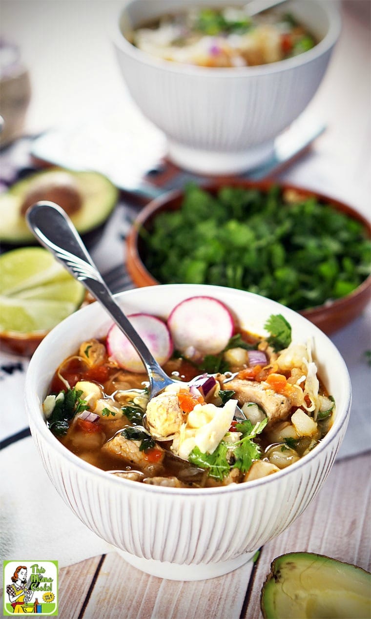 Closeup of spoon in bowl of soup of pozole with a bowl of cilantro in the background.