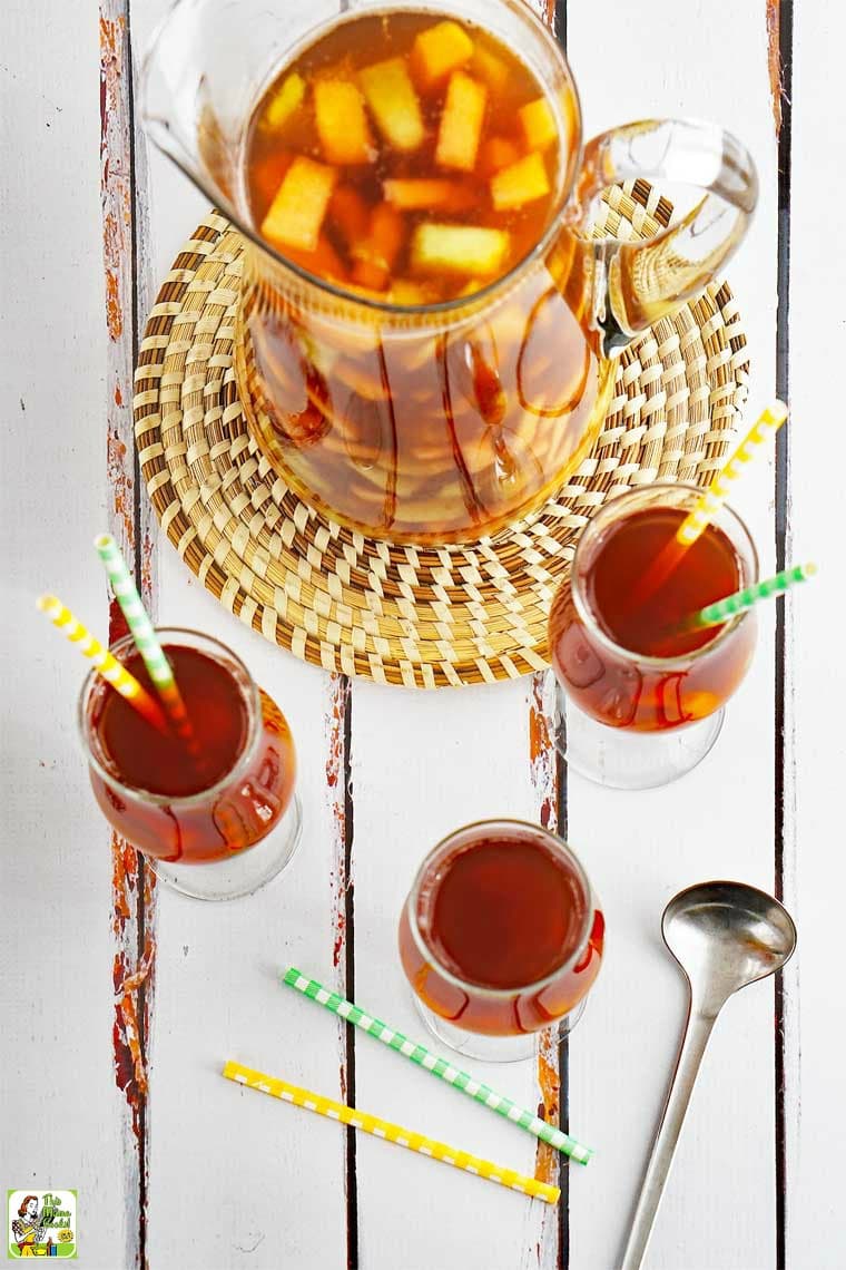 Overhead view of tall glasses and a pitcher of fruit ice tea with yellow and green straws on a woven straw mat with a silver serving spoon.