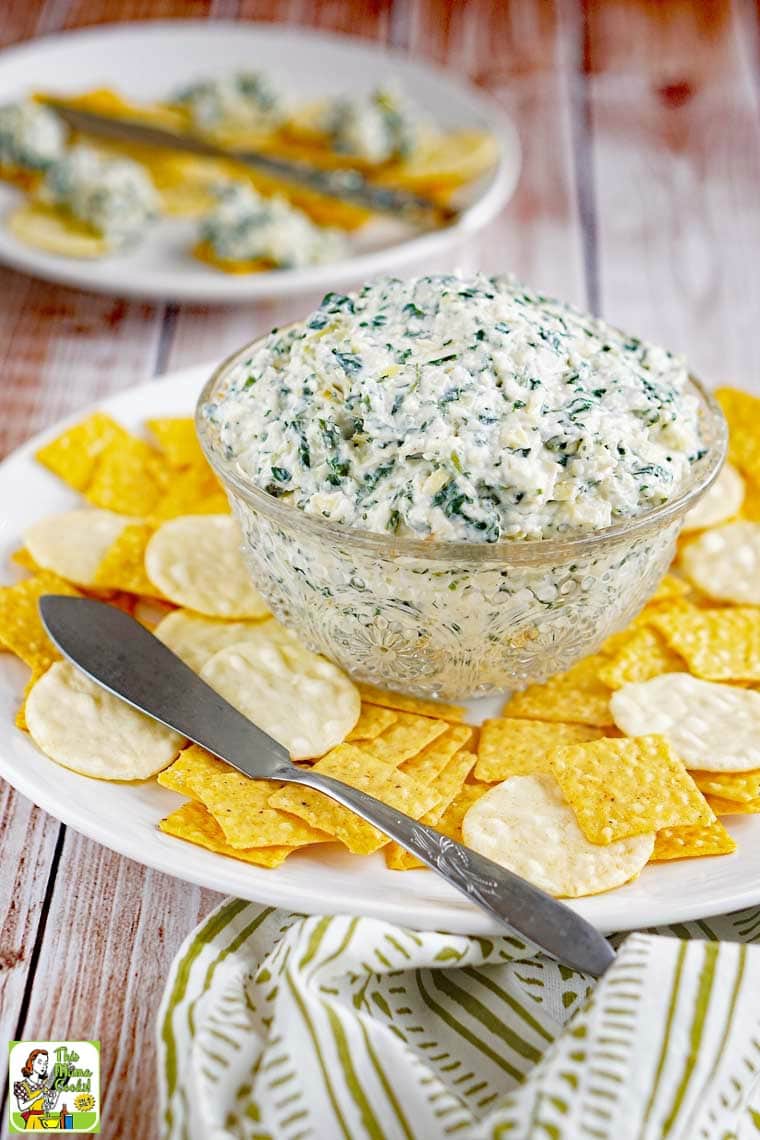 Yellow and white crackers and a bowl of Vegan Spinach Artichoke Dip on a white plate with a serving knife.