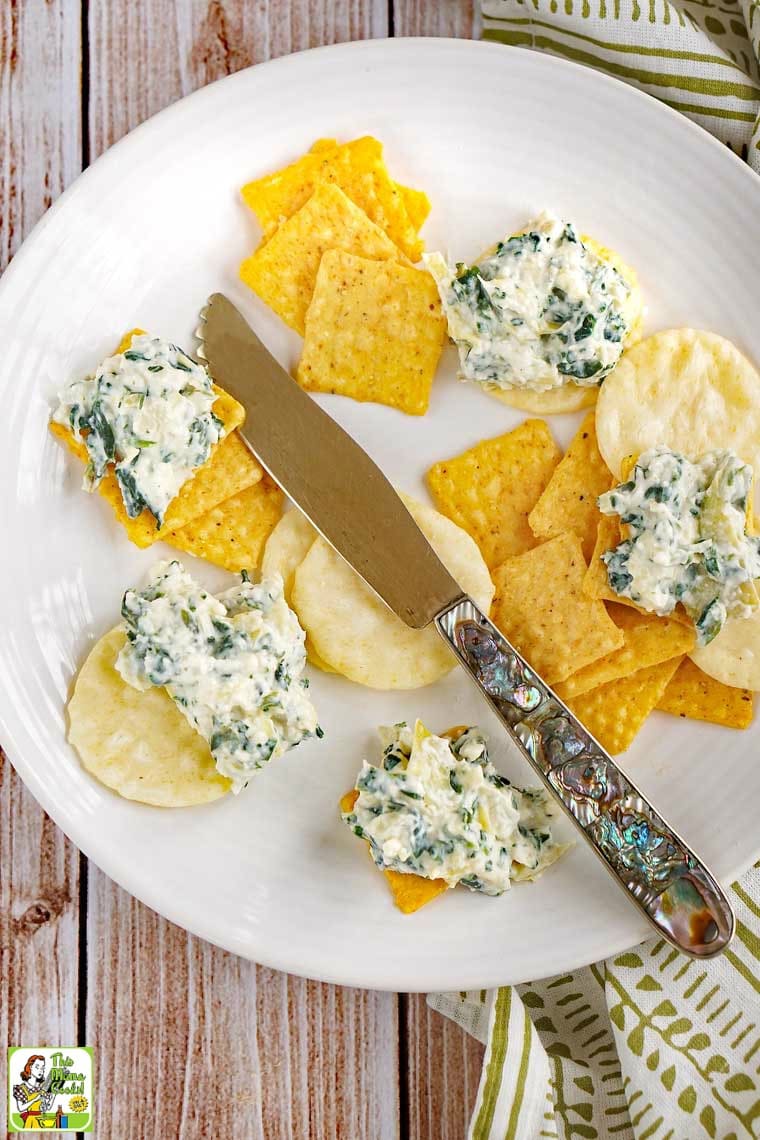 Yellow and white crackers spread with Vegan Spinach Artichoke Dip on a white plate with a serving knife.
