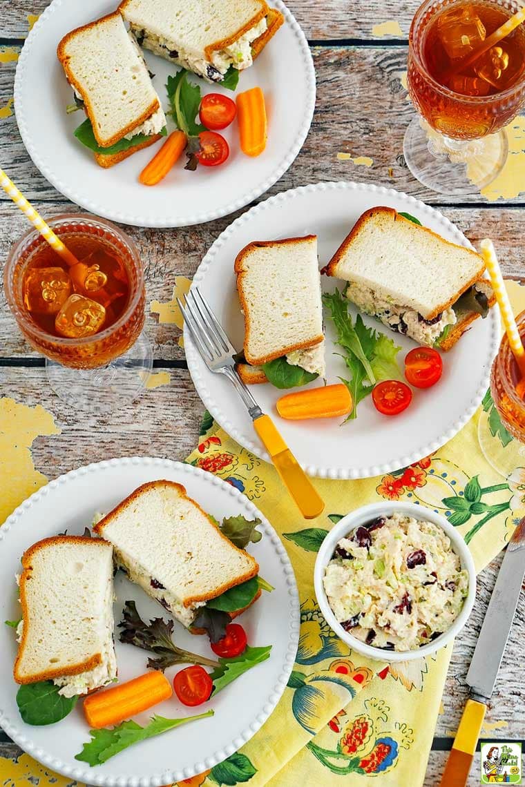 Overhead shot of chicken salad sandwiches on plates with tomato, lettuce, cutlery, and a glasses of iced tea with straws