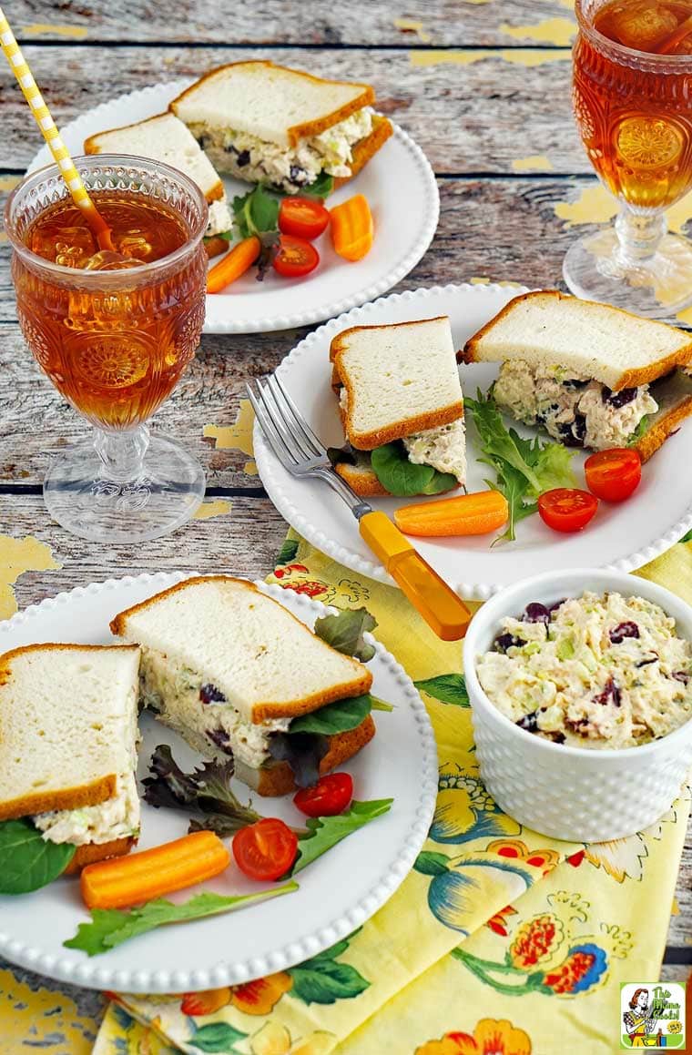 Overhead shot of chicken salad sandwiches on plates with tomato, lettuce, cutlery, and a glasses of iced tea with straws. Served with floral napkins with extra bowls of chicken salad.