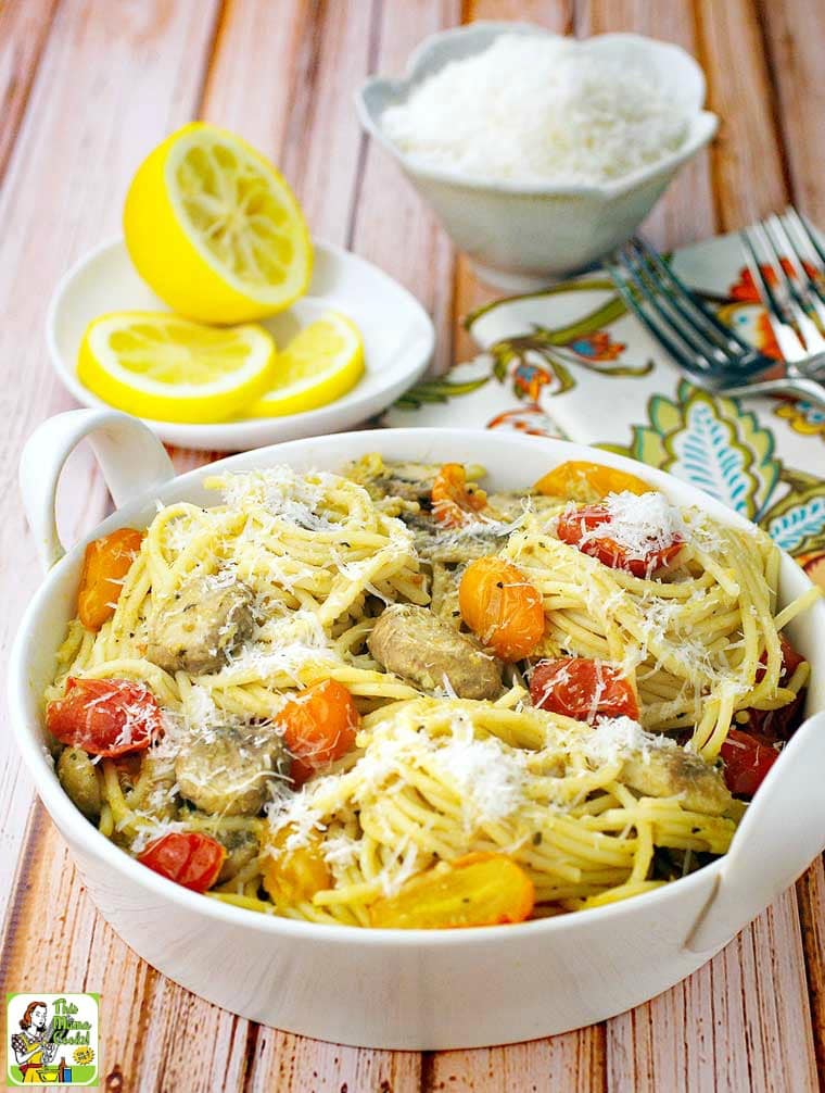 A bowl of spaghetti with crab, pesto, mushrooms, tomatoes and cheese with a plate of lemons, a bowl of cheese, forks and a napkin in the background.