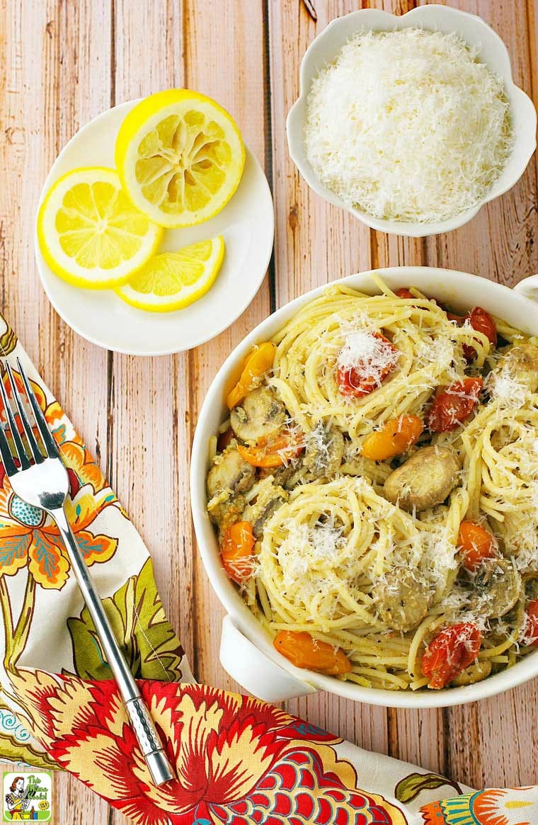 A bowl of Crab Pasta with Pesto, Mushrooms & Tomatoes with a fork, napkin and plate of sliced lemons.