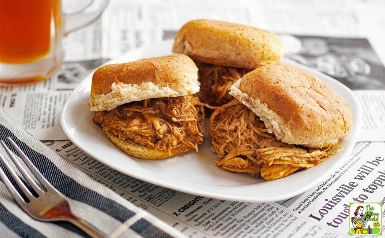 A white plate of Slow Cooker Buffalo Chicken Sliders on a table covered in newspapers with a napkin, fork, and glass of beer.