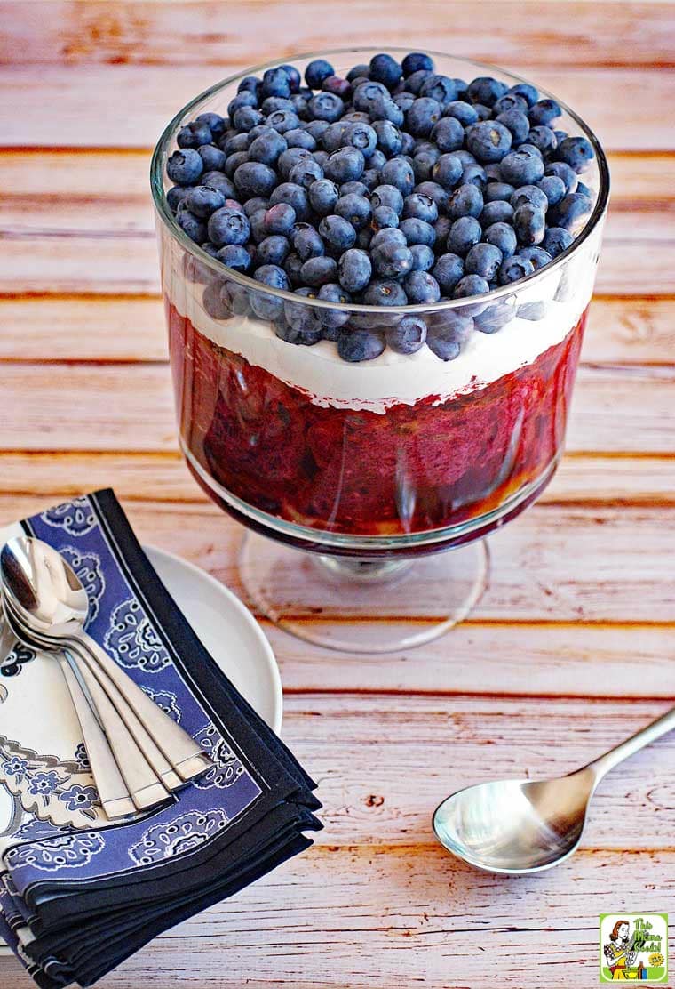 Trifle with blueberries and whipped cream in a glass trifle bowl with blue napkins and serving spoons.