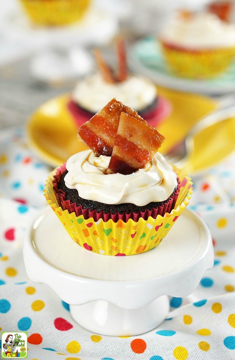 Chocolate bacon cupcakes garnished with candied bacon on a mini cake stand and yellow plate on a polka dot tablecloth.