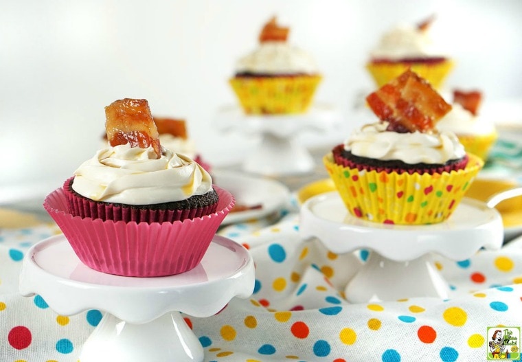 Chocolate cupcakes with vanilla frosting garnished with candied bacon on a mini cake stands on a polka dot tablecloth.