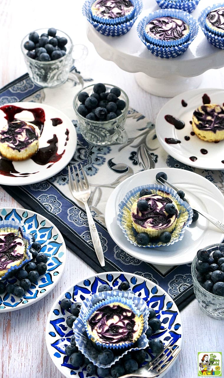 Plates of mini blueberry cheesecake cupcakes with forks, cups of blueberries on a blue and white napkin. 