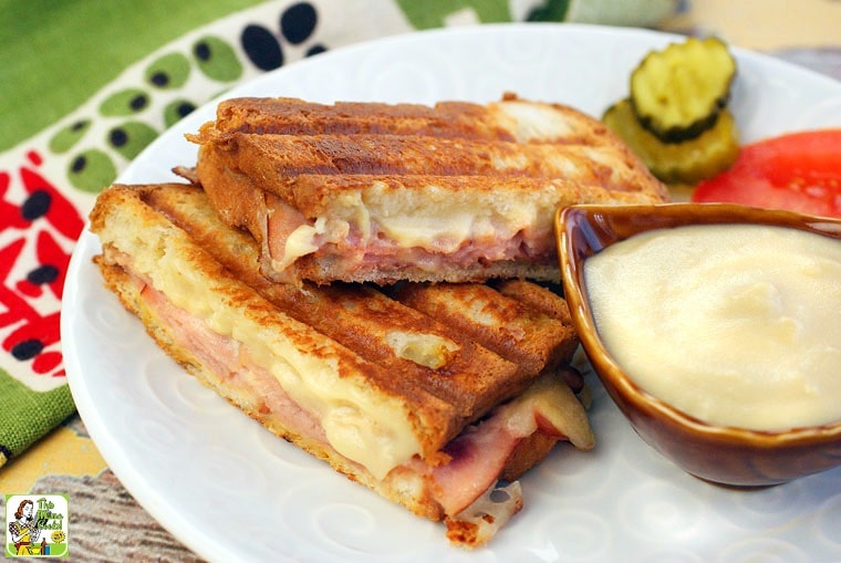 A plate of Croque Monsieurs with a dish of Honey Bechamel Sauce, pickles, and tomato.