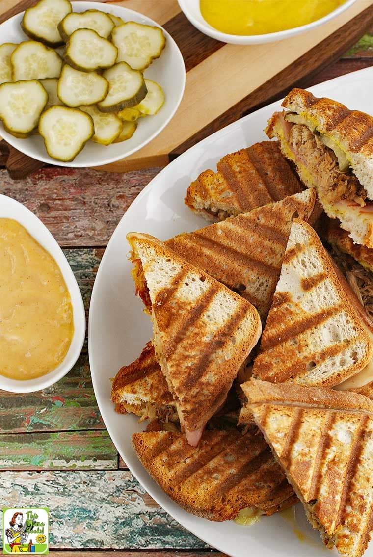 Overhead shot of a pile of Cuban panini sandwiches on a white platter with dishes of pickles, mustard and dressing.