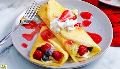 Two gluten free crepes filled with creme and berries covered in whipped cream and strawberry sauce on a grey plate with a fork.