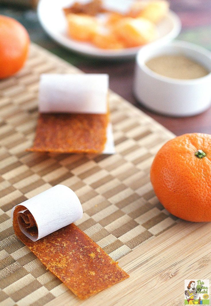 Closeup of Orange Fruit Leather rollups and oranges on a wooden cutting board with spices and orange slices in the background.