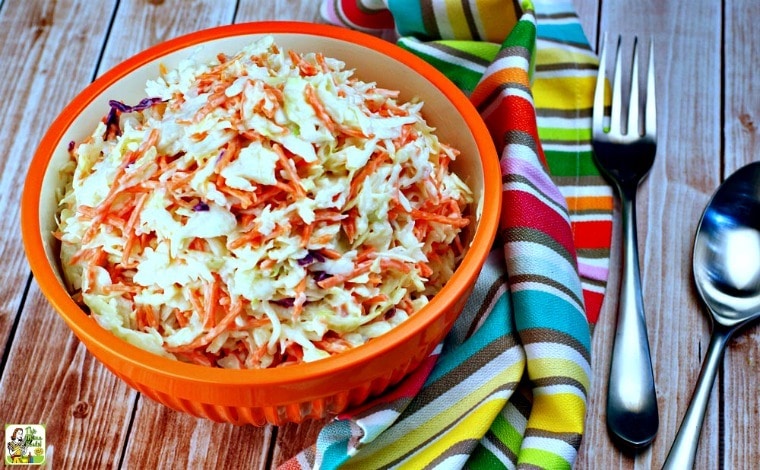 An orange bowl of Easy Coleslaw with striped kitchen towel and serving fork and spoon.