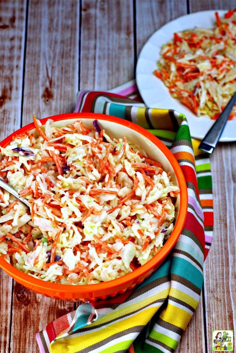 An orange bowl and plate of coleslaw with striped towel and serving fork and spoon.