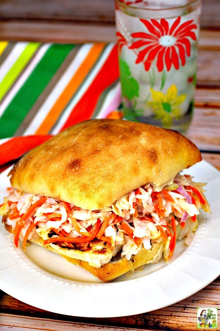 Coleslaw on grilled chicken sandwich on a plate with glass of water and striped napkin.