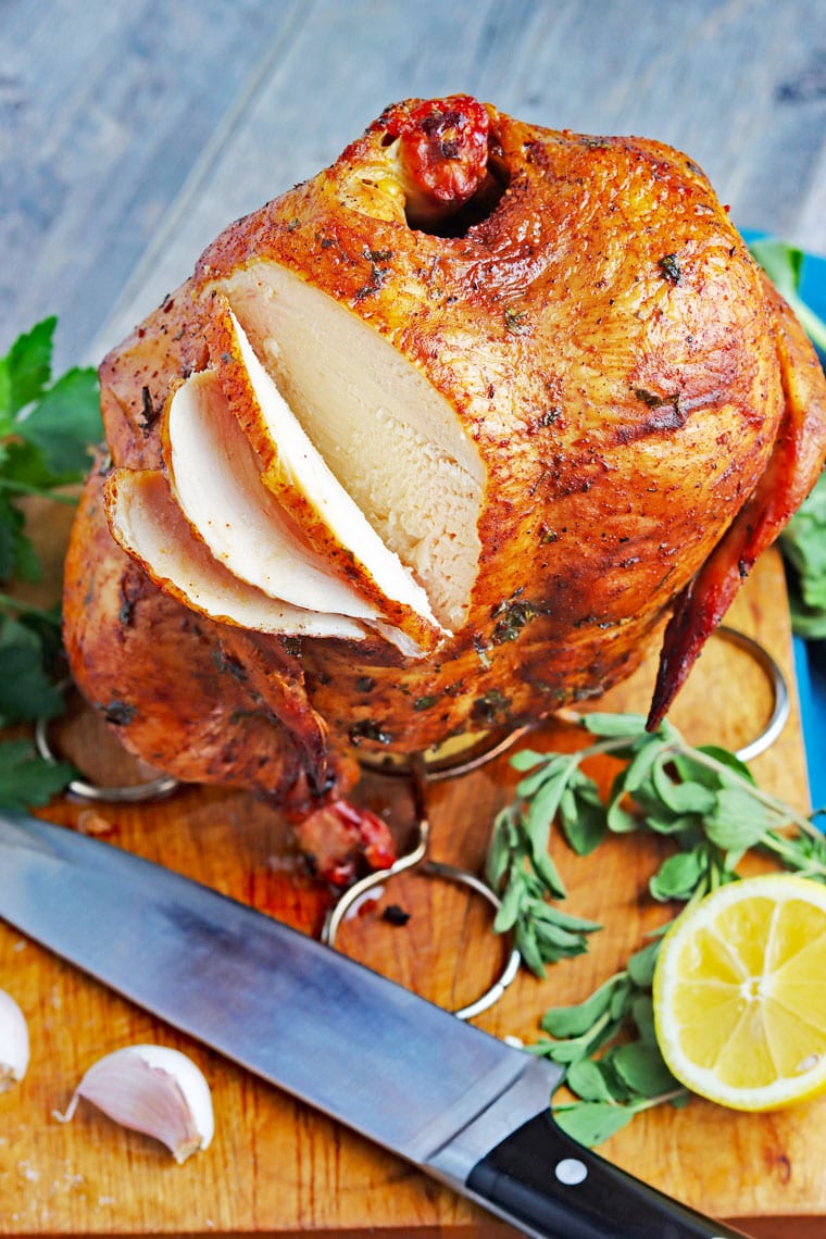 Sliced smoked beer can chicken on a wooden cutting board with a knife, lemon and fresh herbs.