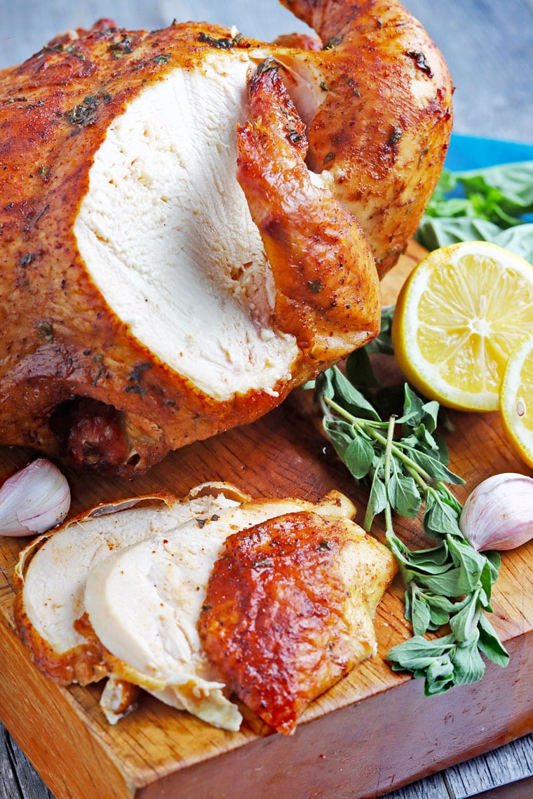 Sliced roasted chicken with pieces of chicken on a wooden cutting board with herbs and sliced lemon and garlic.
