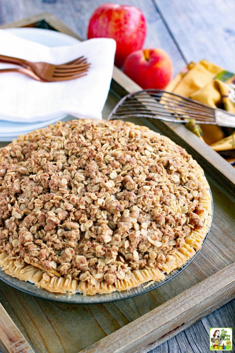 A peach pie with crumb crust on a wooden tray with plates, napkins, forks, apples, and a pie server.
