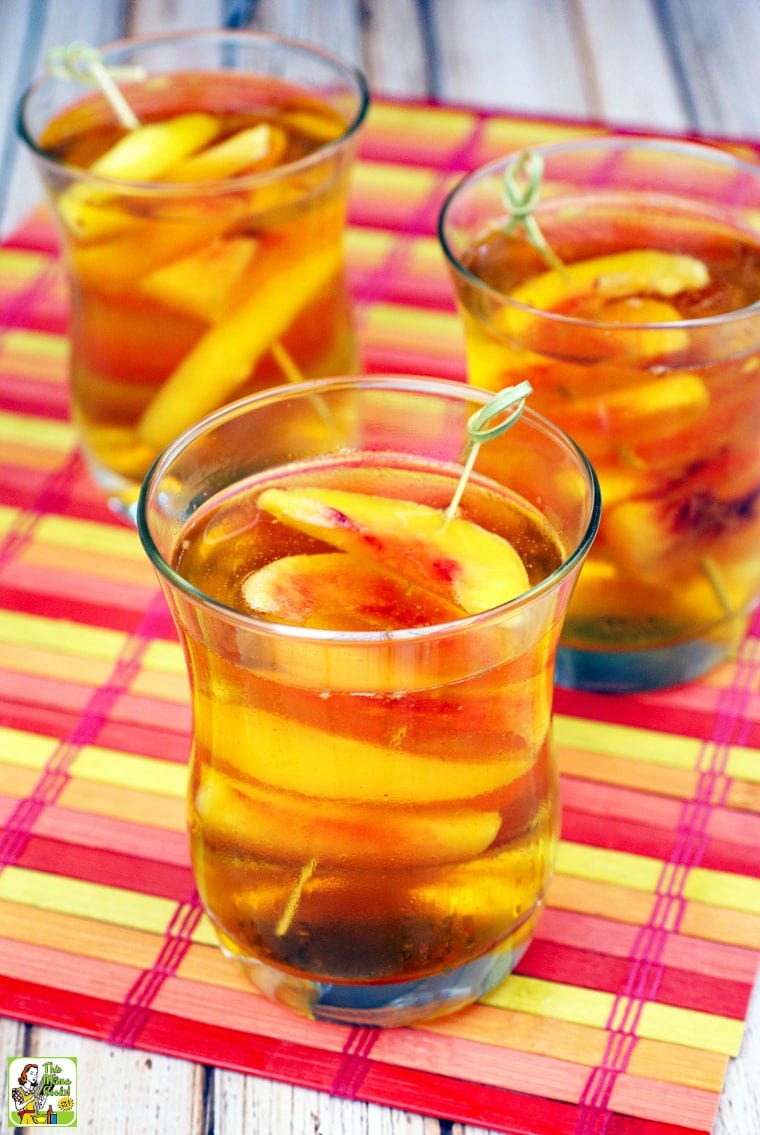 3 glasses of Peach Whiskey Cocktail with slices of peaches on bamboo skewers on a colorful bamboo placemat.