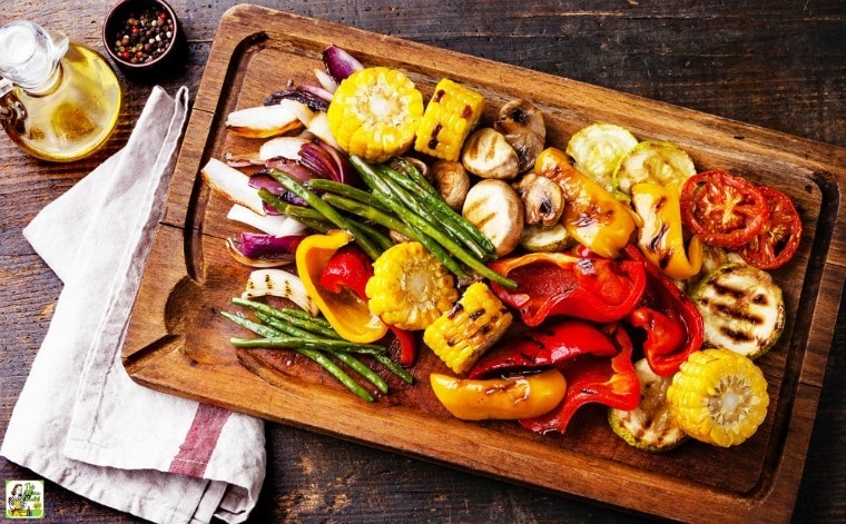 A wooden cutting board of Roasted Marinated Vegetables with bottle of oil, ramekin of peppercorns, and kitchen towel.