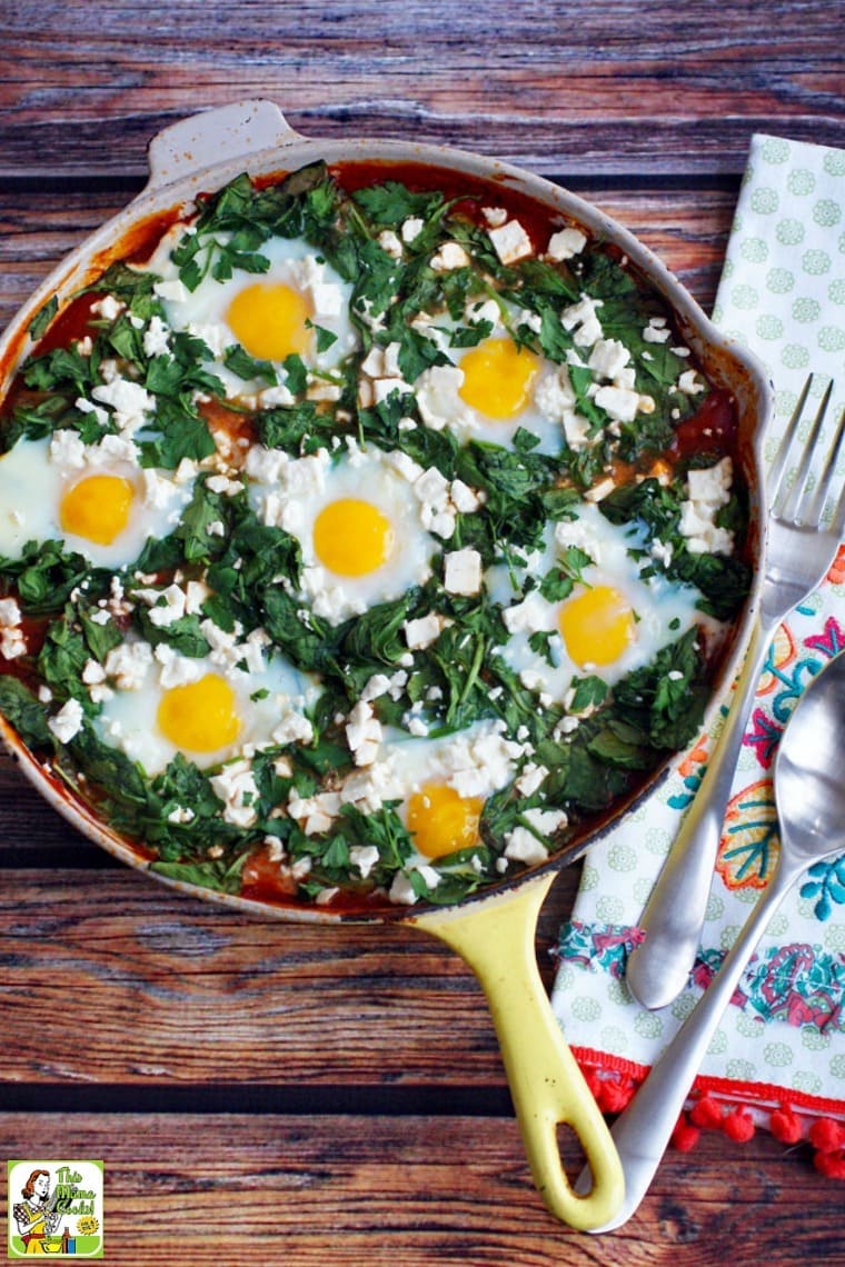 Overhead view of shakshuka with feta in a yellow skillet with silver serving spoon and fork on an embroidered tea towel.