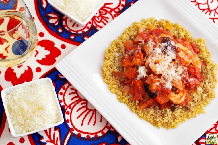 A bowl of shrimp puttanesca on quinoa on a red and blue tablecloth with a glass of wine and two small bowls of shredded cheese.