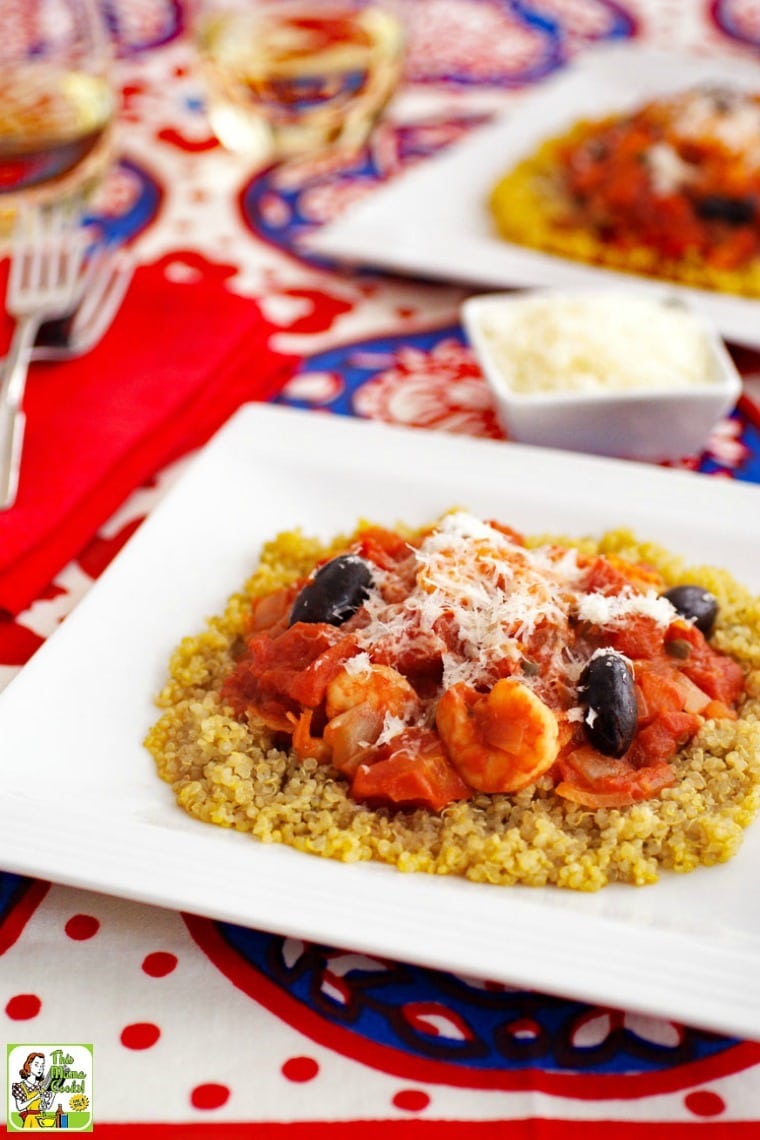 Bowls of shrimp puttanesca on a colorful tablecloth, glasses of white wine, red napkin and forks, and small bowls of shredded cheese.