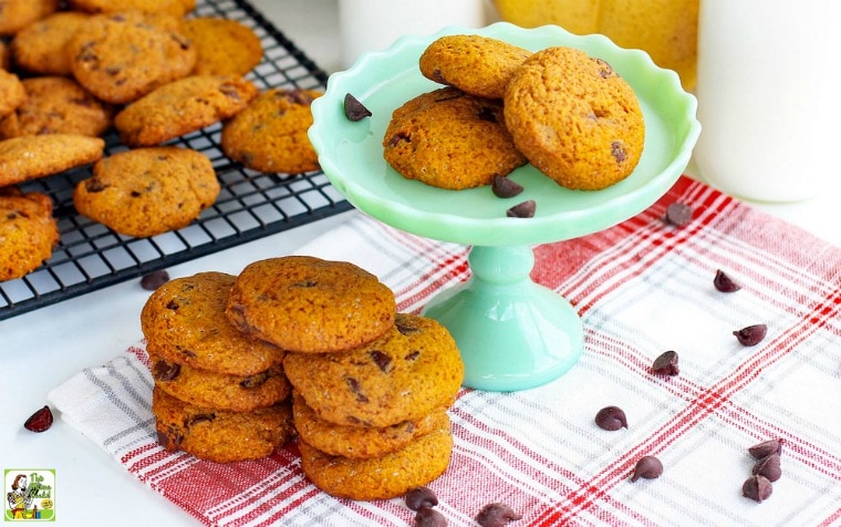 Stacks of chocolate cookies on a green cake stand, baking rack, and a red and white napkin with pumpkins and milk bottles in the background.