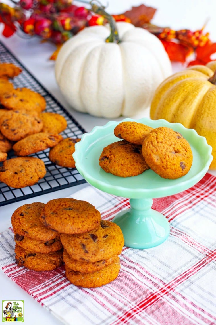 Stacks of chocolate chip cookies on a green cake stand, baking rack, and a red and white napkin with pumpkins in the background.