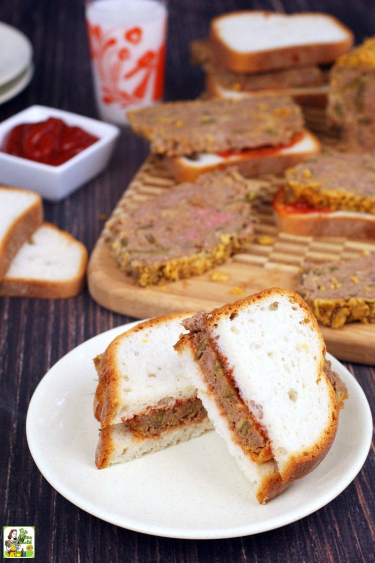 A meatloaf sandwich on a plate with more sandwiches and condiments on a cutting board in the background.