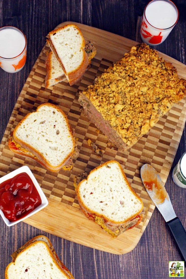 Overhead shot of meatloaf sandwiches, meatloaf, ketchup, and glasses of milk on a wooden cutting board.