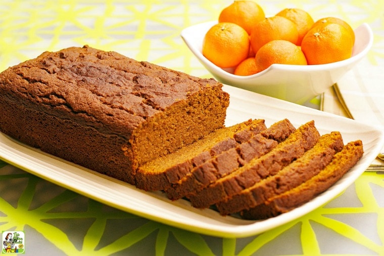 A loaf of  Pumpkin Bread sliced up on a white plate with a bowl of tangarines in the background.