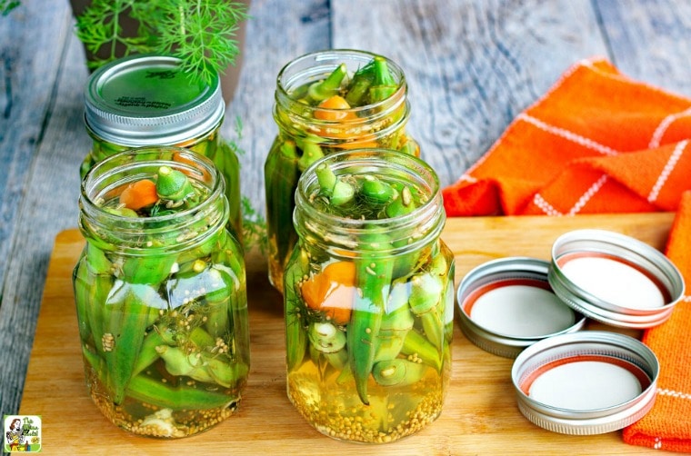 Jars of Refrigerator Pickled Okra with lids on a wooden cutting board with orange cloth napkins.