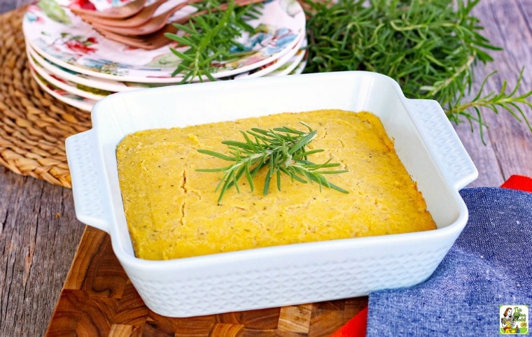 A white baking dish of Vegan Cornbread Recipe with fresh rosemary, plates, and forks with a woven mat, cutting board, and cloth napkins.