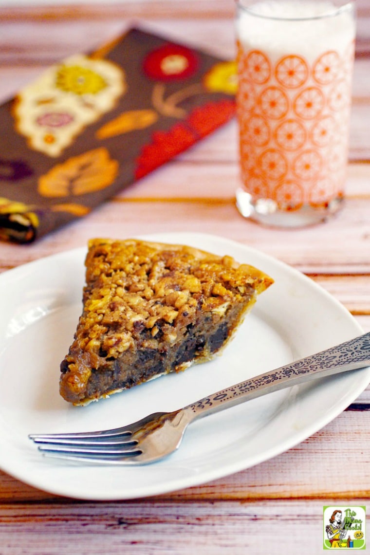 A slice of gluten free chocolate pecan pie on a white plate with a silver fork. Orange vintage glass of  milk and brown napkin in the background.