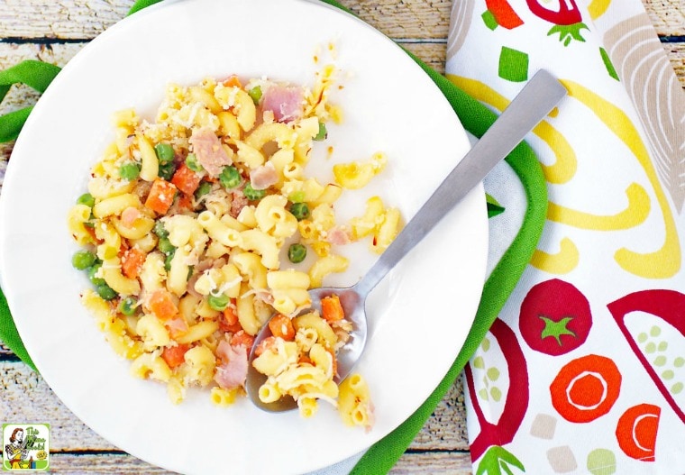 A plate of Mac & Cheese Casserole with Peas, Carrots & Ham on an oven mitt with vegetable patterned kitchen towel.