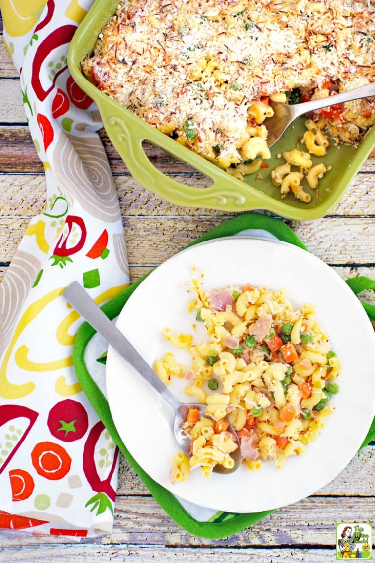 A plate of mac and cheese with peas, carrots & ham in a green casserole dish.