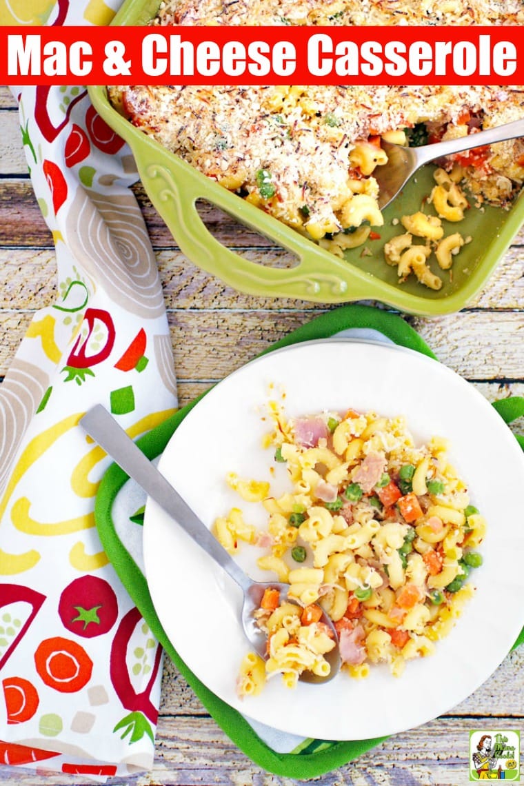 A plate of mac and cheese with peas, carrots & ham in a green casserole dish and a veggie patterned dish towel.