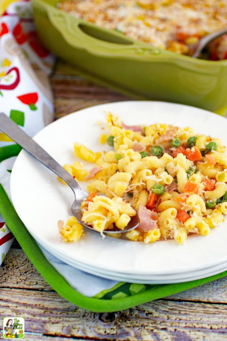 A white plate of mac and cheese with peas, carrots & ham.