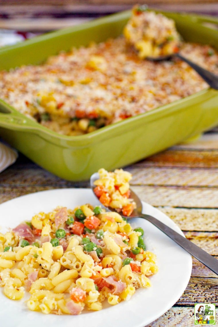 A plate of mac and cheese with peas, carrots & ham in a green casserole dish.