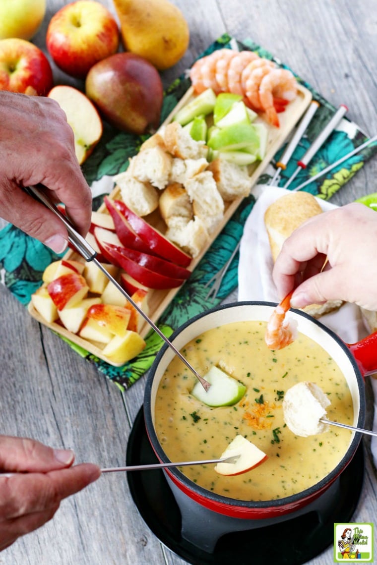 Hands dipping fruit into brie cheese fondue recipe in red pot. Fruit, bread, shrimp and other fondue dippers on tray.