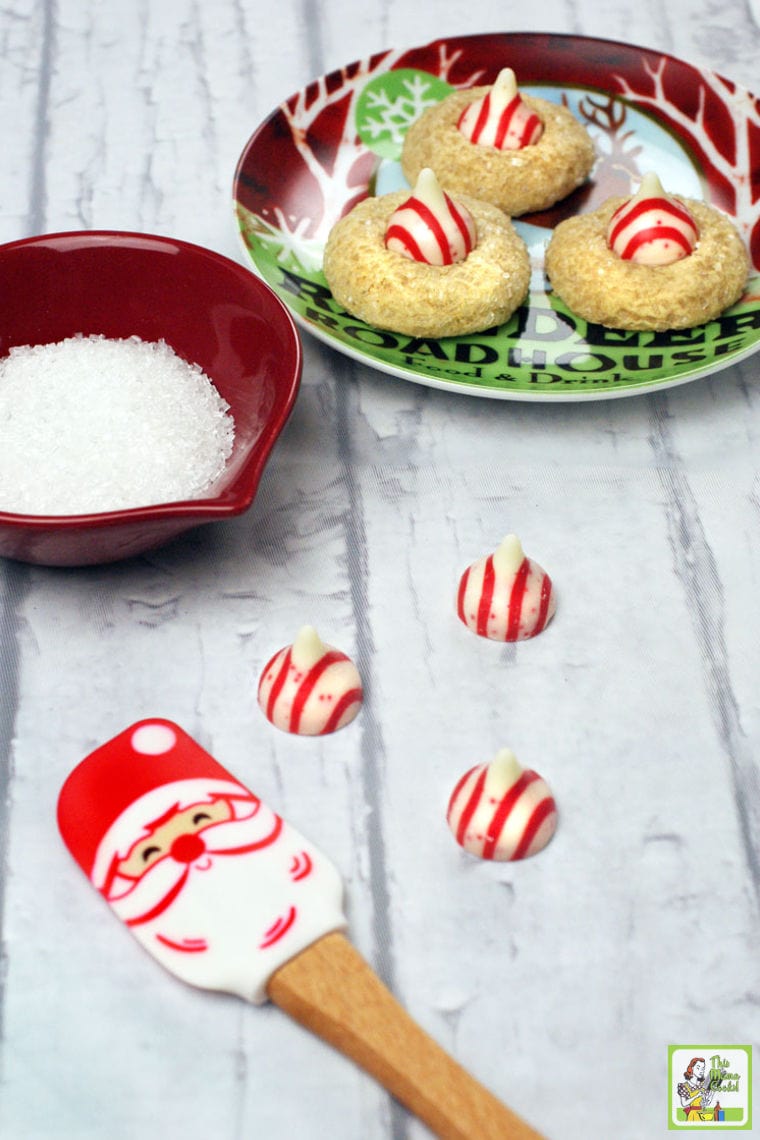 A Christmas plate of thumbprint cookies with striped Hersey Kisses, with bowl of decorative sugar, Hersey's kisses, and Santa spatula.