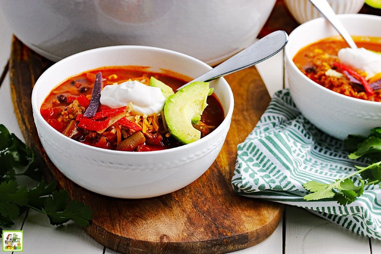 White bowls of 7 Can Taco Soup with dollops of sour cream and a slice of avocado on a wooden cutting board and a green and white napkin.