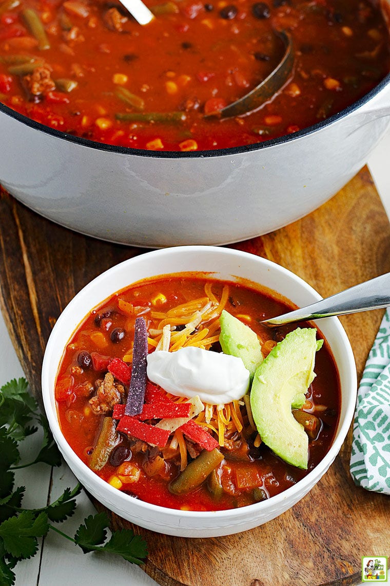 Overhead shot of a cooking pot and bowl of taco soup with spoons on a wooden cutting board with a napkin and bunch of cilantro.