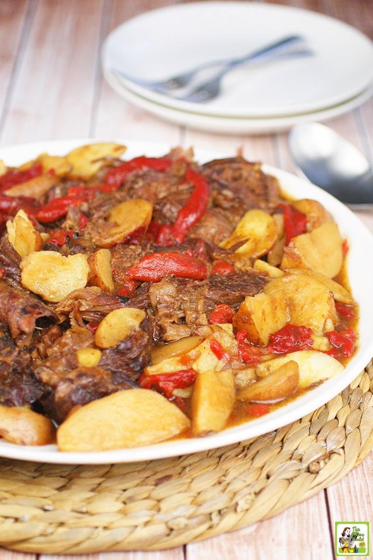 A plate of pot roast with red peppers and potato wedges on a straw mat with plates and forks in the background.