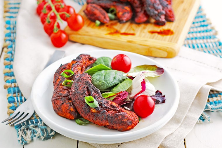 A plate of copycat Popeyes blackened chicken tenders with salad and tomatoes with wooden cuttin board and more chicken in the background.